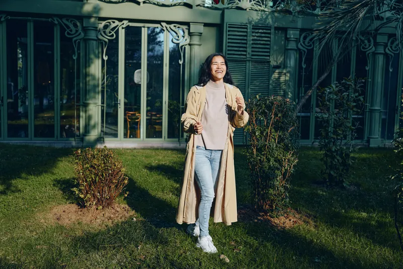 Woman in a long beige coat standing on a green lawn outside a contemporary glass house, casual chic style, sunlight highlights fabric texture and relaxed mood in an outdoor fashion scene.