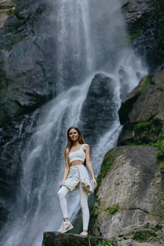 young woman standing near waterfall in activewear with jacket tied around waist, surrounded by rocky nature landscape and lush greenery, outdoor adventure vibe