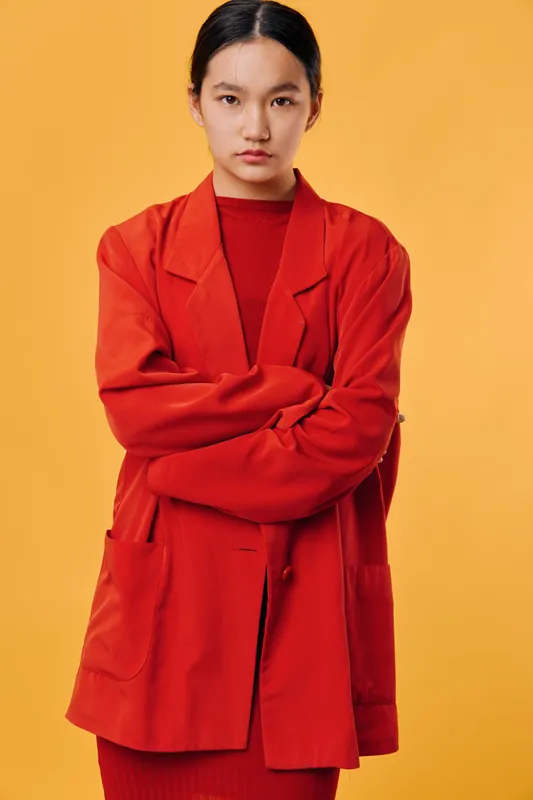 Young woman with serious expression wearing red oversized blazer and matching dress standing with crossed arms on orange background for fashion style and confidence.