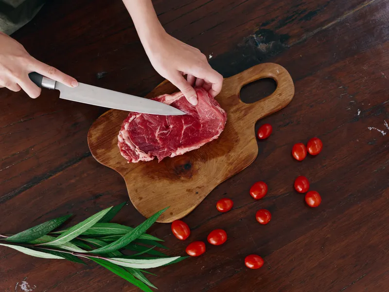 Женщина с ножом в руке нарезает свежее мясо для стейка, предназначенное для запекания на кухне