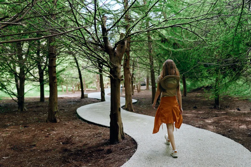 Woman walking on a winding path through a dense forest with a backpack and tall trees, wearing an orange skirt. Solitary outdoor journey on a gravel walkway in summer nature exploration.