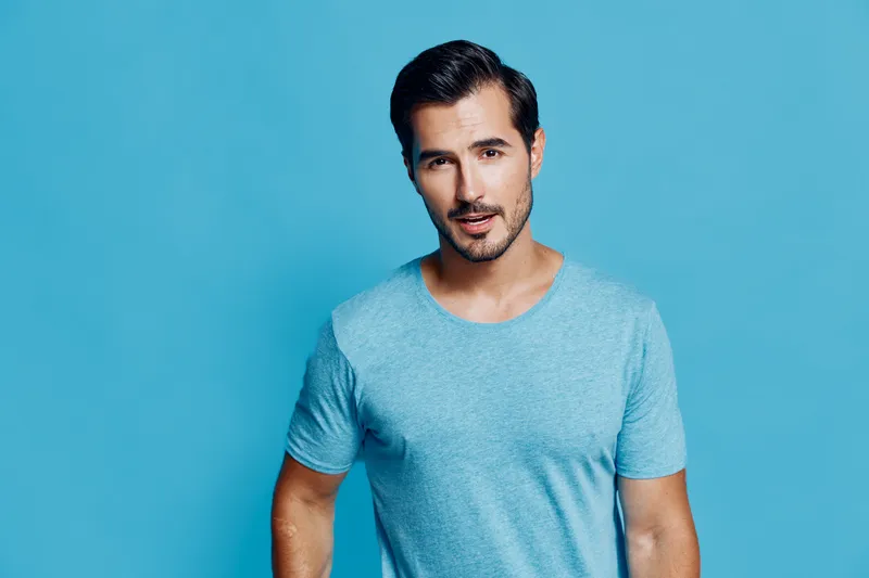 Handsome young smiling man wearing a light blue t-shirt stands against a vibrant blue background, looking directly at the camera with a friendly expression.