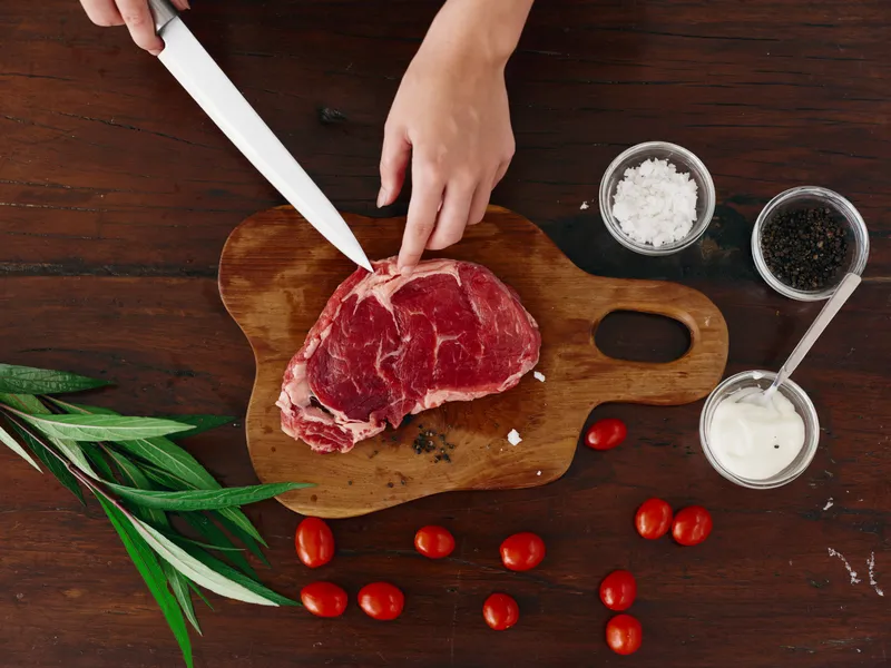 Женщина с ножом в руке нарезает свежее стейковое мясо для запекания на кухне