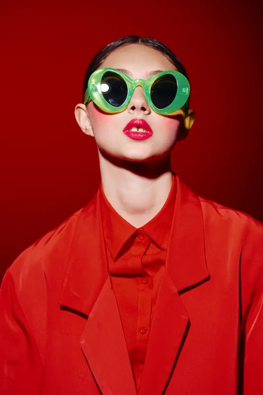Fashionable young woman in bright red jacket poses with oversized translucent green sunglasses on vivid red background. Concept for bold beauty, creative style, and unique fashion editorial.