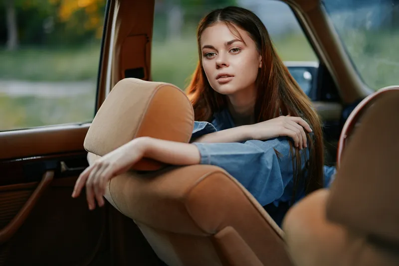 Young woman with long hair in denim attire sitting inside a vintage car, exuding calm emotions in a nature-inspired background.