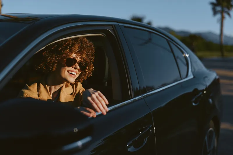 Cheerful woman leaning out of a dark car window, sunlit outdoors, curly hair, retro film tones, relaxed posture, natural colors, candid moment on a sunny road