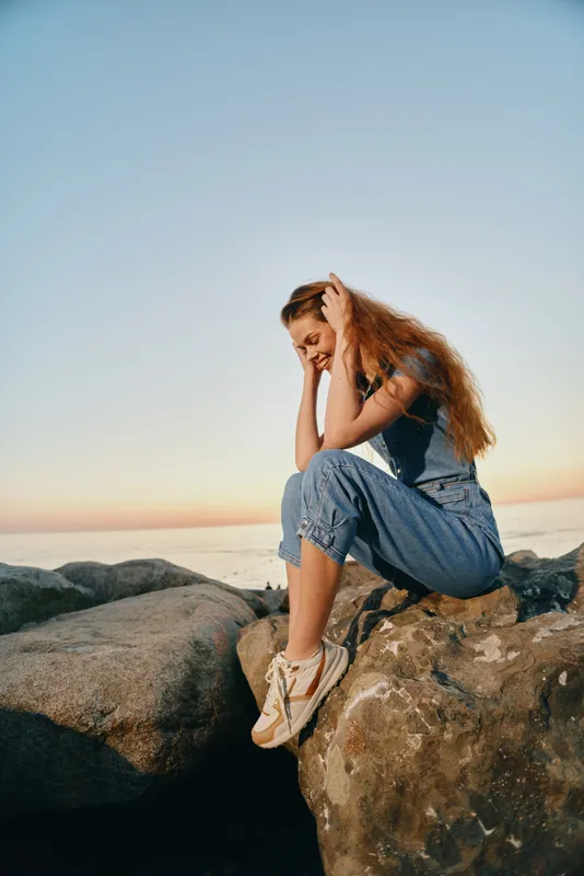 lifestyle woman sitting on rocks by the sea in modern boho-western denim street style with natural light and film color tones at sunset