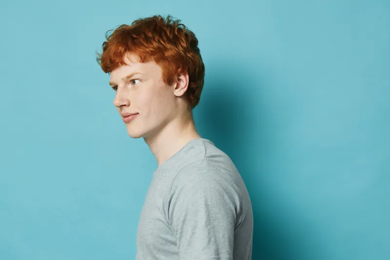 Young man with red hair and pale skin wearing a gray long sleeve shirt standing against blue background, side view portrait with casual expression and natural light.