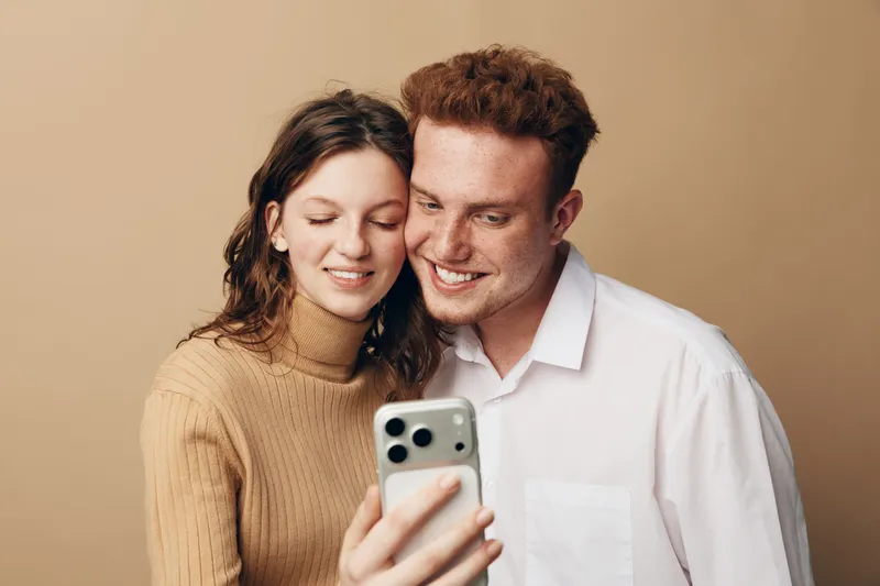 Young couple with curly hair smiling and looking at smartphone screen together. They wear casual clothes and stand close on beige background. Concept of friendship, technology, and happiness.