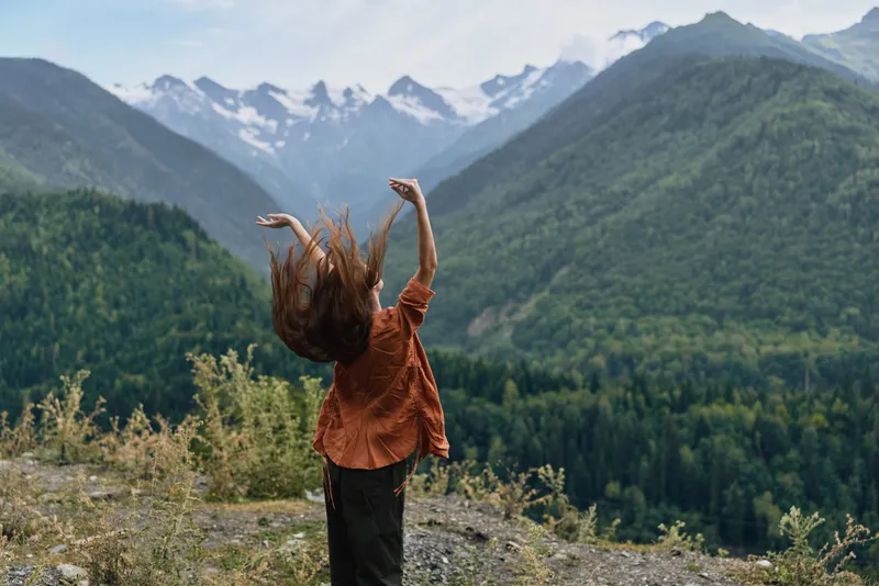 woman person mountain landscape freedom nature authenticity inclusivity from back on rocky viewpoint overlooking peaks and forest arms raised in joyful outdoor moment