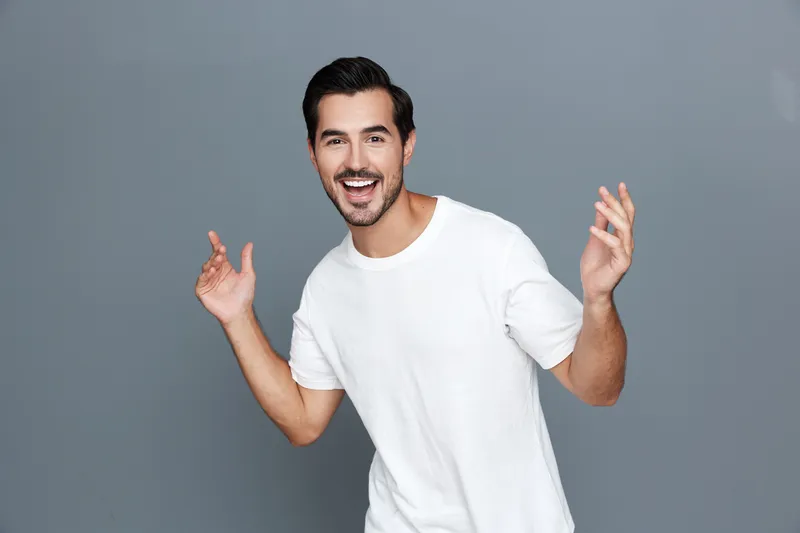 Full body smiling happy cheerful young man wearing a casual white t-shirt poses confidently with his hands raised in joy against a plain gray background.