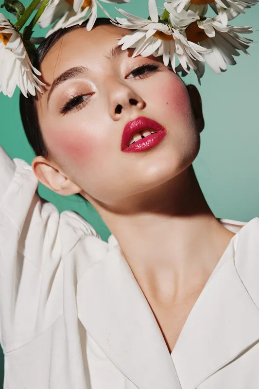 Close-up portrait of a fashion woman wearing glossy red lipstick and pink blush with a delicate flower crown on green background, showcasing creative beauty concept.