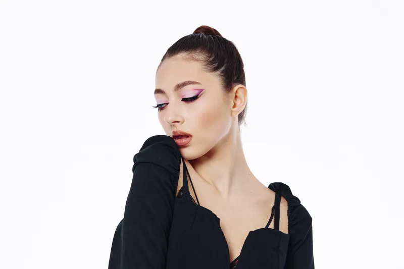 Young elegant woman with high ponytail wearing black clothing, leggings and vivid pink eyeliner makeup on eyelids. Studio portrait on white background monochrome concept beauty and fashion.