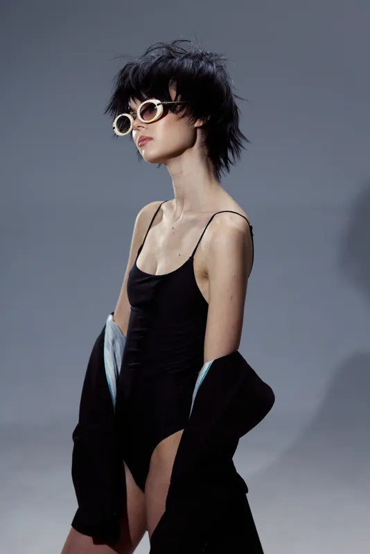 Stylish woman wearing black swimsuit and sunglasses posing against gray background