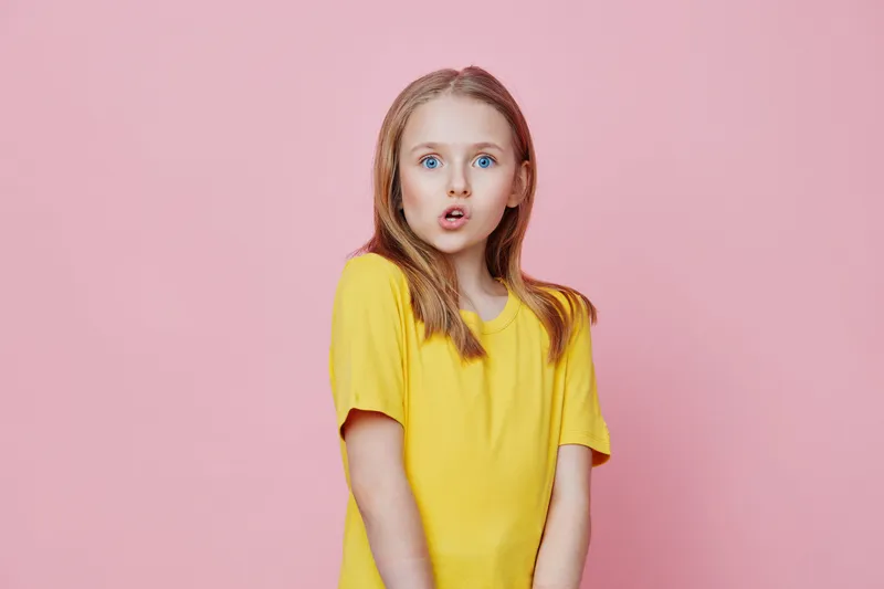Young girl with surprised expression wearing a yellow shirt against a pink background, capturing joyful childhood moments and vibrant emotions in a playful setting