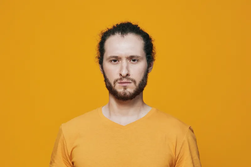 Young man with curly hair wearing an orange shirt, posing against a vibrant yellow background, expressing confidence and determination, perfect for emotional branding