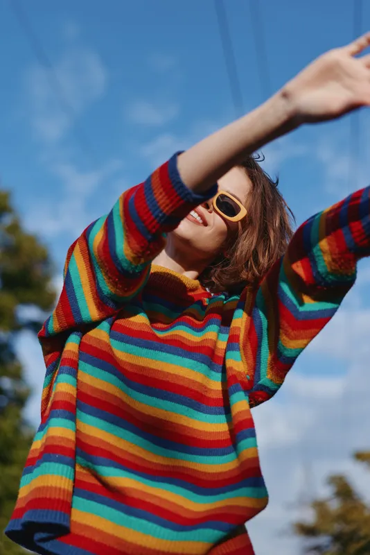 Woman in rainbow striped sweater and sunglasses smiling outdoors with arms raised against blue sky, joyful portrait, casual fashion, bright colors and sunny mood, carefree movement.