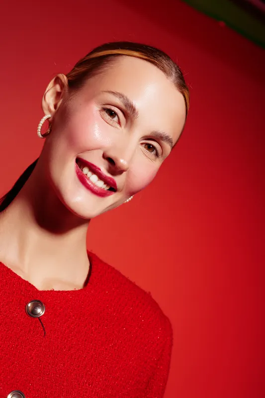 Smiling woman with red hair and bright lipstick wears red sweater against red background. Close-up portrait with hoop earrings and natural skin glow. Concept of beauty, fashion, and confidence.
