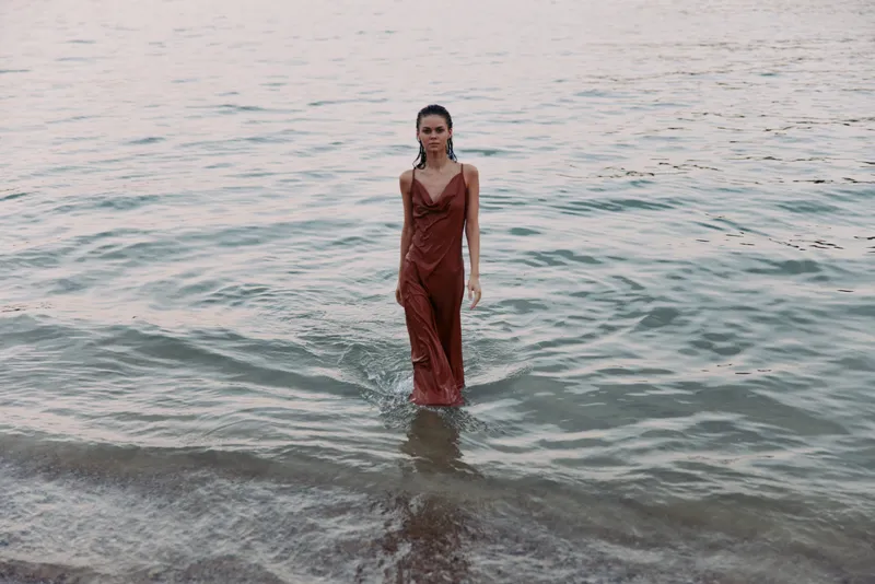 Woman stands in shallow sea water wearing a flowing red dress, creating a bold focal point as gentle waves lap around her and soft light glints on the surface.