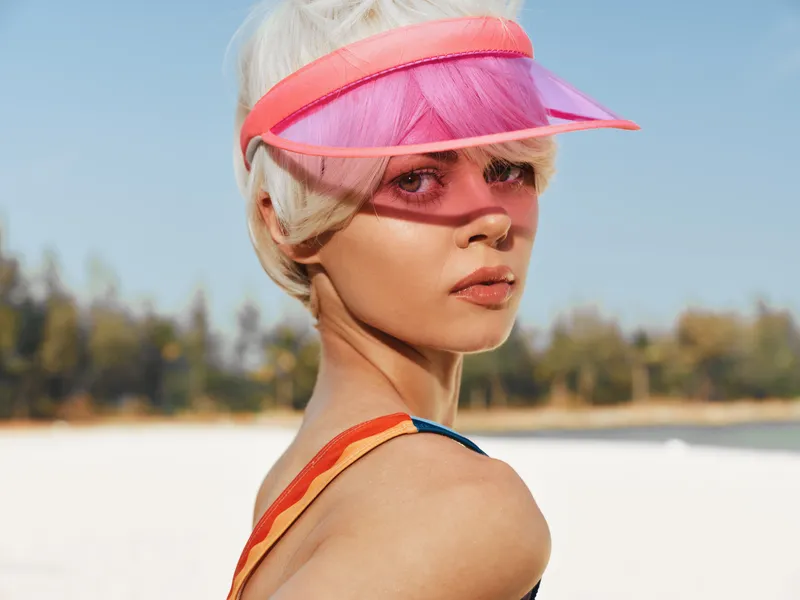 Portrait of a stylish woman on a sunny beach wearing a pink visor and sheer veil, sunlit skin and windswept hair create a bold fashion moment.