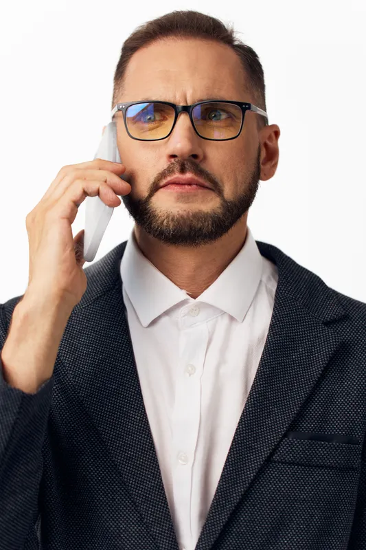 A professional man in business attire sits against an isolated blue background, showing focused emotion while speaking on a mobile phone during a work call.