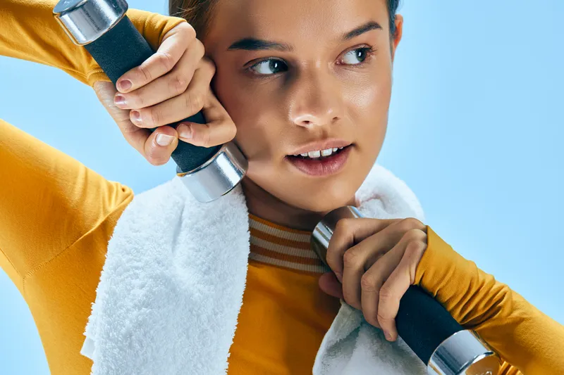 Woman fitness athlete in bright orange top lifting dumbbells with a towel around her neck, concentrating on form during a dynamic workout in a bright studio.
