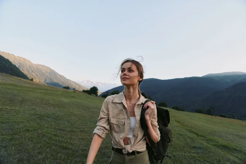 Hiking woman moves through a grassy valley with a mountain backdrop, wearing a beige shirt and backpack, looking up with a hopeful, thoughtful expression.