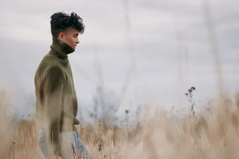 Young man in a textured sweater walking through tall dry grass, photographed in a candid, natural moment that emphasizes genuine presence and visual realism in soft overcast light.