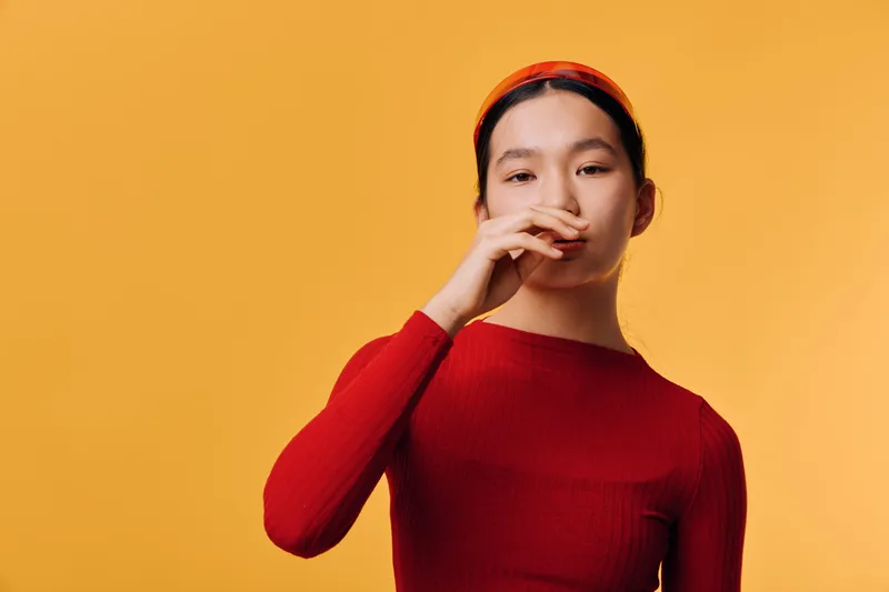 Young woman in red sweater and headband covering mouth with hand standing on orange background. Studio portrait of female model with thoughtful or shy expression.