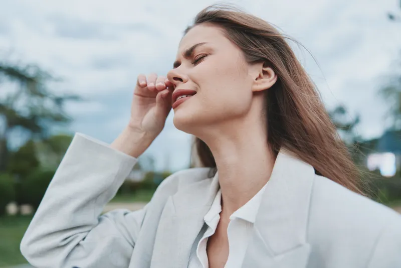 Woman in light gray jacket feeling stressed outdoors with closed eyes and natural background, emotional expression reflecting psychological tension and fatigue