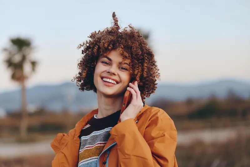Happy woman lifestyle outdoors, curly hair woman smiles while talking on phone call, warm jacket, natural landscape background, casual travel portrait, cheerful communication.