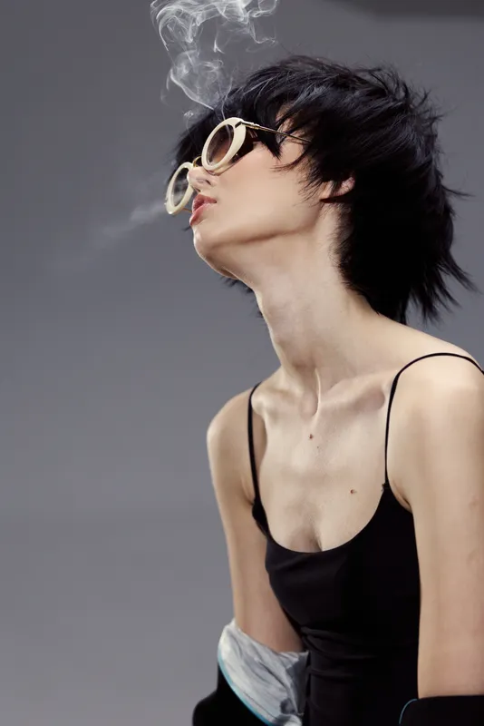 Stylish woman exhaling smoke with sunglasses in a modern fashion pose, minimalistic background and moody lighting.
