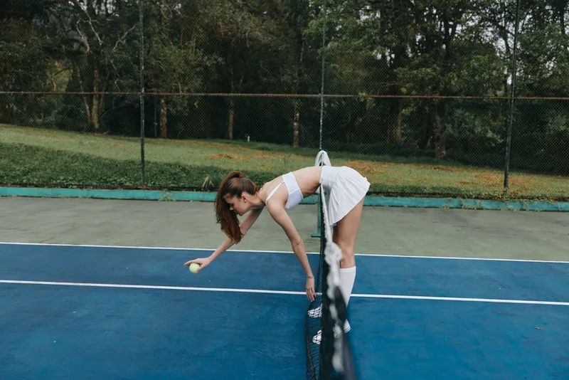 Young woman in sporty outfit preparing to serve on tennis court, showcasing focus and determination with a green nature background and vibrant blue court