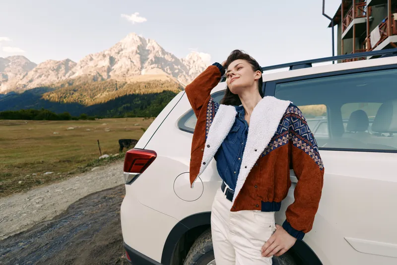 Woman leaning on car beside mountains during travel roadtrip, portrait of relaxed traveler in cozy jacket and nature scenery, casual shorthair style and scenic outdoor adventure.