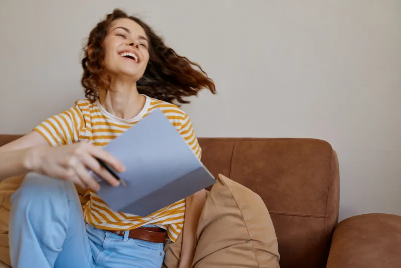 Woman lifestyle casual, relaxed indoors on a sofa, holding papers with a cheerful smile, warm natural light, cozy interior, candid moment showcasing everyday home life and comfort
