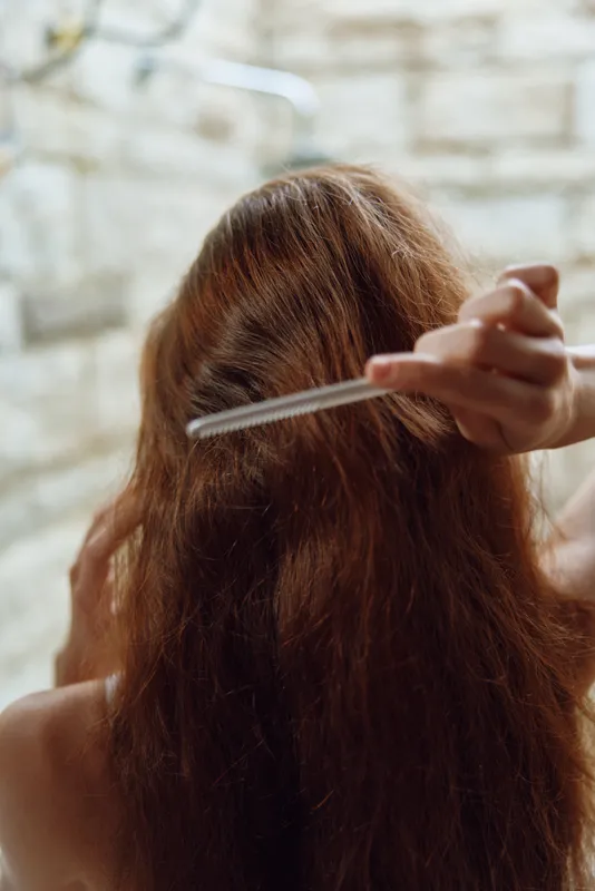 Woman combing long brown hair with a fine-tooth comb in a cozy, softly lit indoor bathroom setting, showing detailed hair texture and natural beauty.