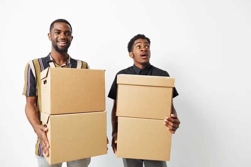 Two smiling black men carrying cardboard boxes, displaying a sense of excitement and teamwork while preparing for a move or delivery. Perfect for conveyance roles.