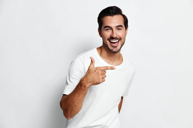 Smiling young man in a casual white t-shirt posing against a plain light background, pointing with his right hand, exuding confidence and joy.