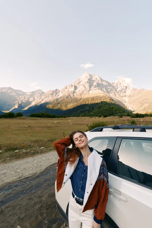 Woman by car in mountains and nature, travel portrait on road and meadow. Young woman leaning on SUV, relaxed pose, scenic alpine landscape and sunny outdoor adventure.