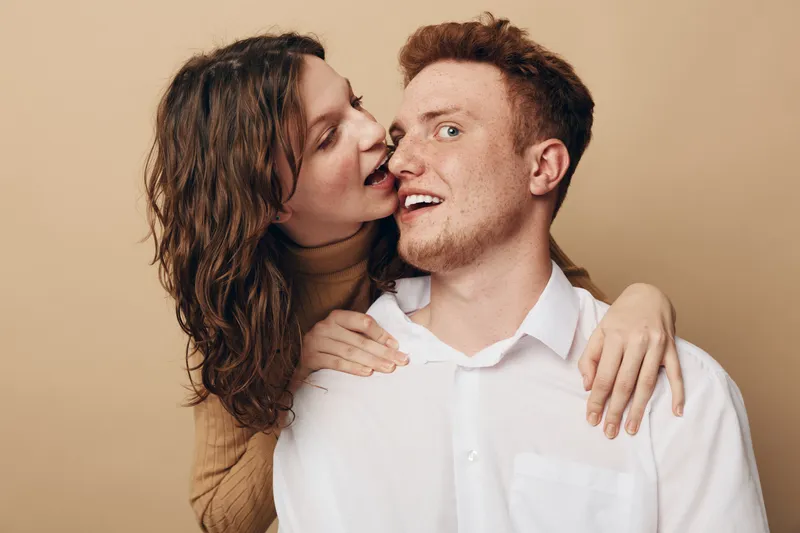 Happy young couple in casual clothes share intimate moment on beige background. Woman embraces man from behind with playful smile. Concept of love, affection, and relationship.
