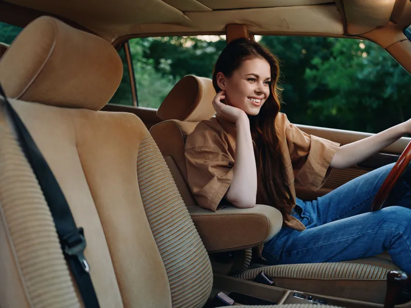 Young woman smiling in a vintage car, wearing a casual brown outfit, showcasing joy and relaxation in a lush green setting, evoking nostalgia and warmth.