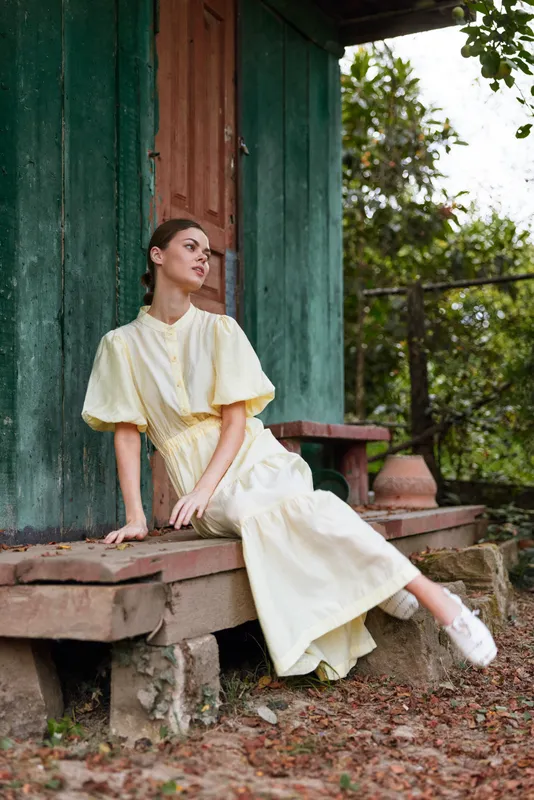 Woman in a flowing yellow dress sits gracefully on wooden steps beside a rustic doorway, outdoor setting, soft natural light, calm mood, fashion elegance in a rural atmosphere.