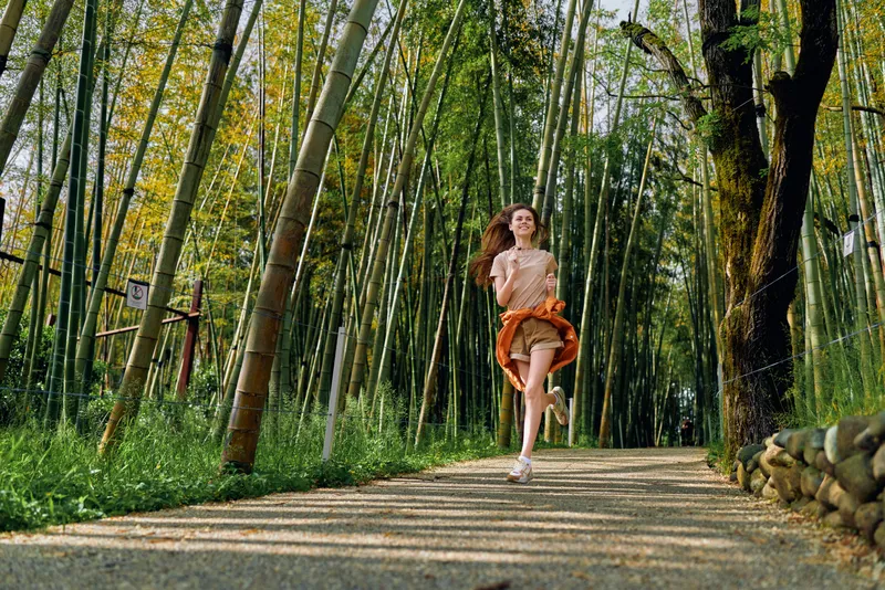 Woman running through a bamboo forest path, smiling and jogging outdoors in a casual skirt and sneakers. Motion and sunlight on green trees and gravel trail create a carefree active portrait in