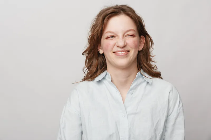 Young woman with wavy brown hair wearing a white shirt makes a funny face with a scrunched nose and closed eyes on a plain light background. Concept of humor and candid expression.