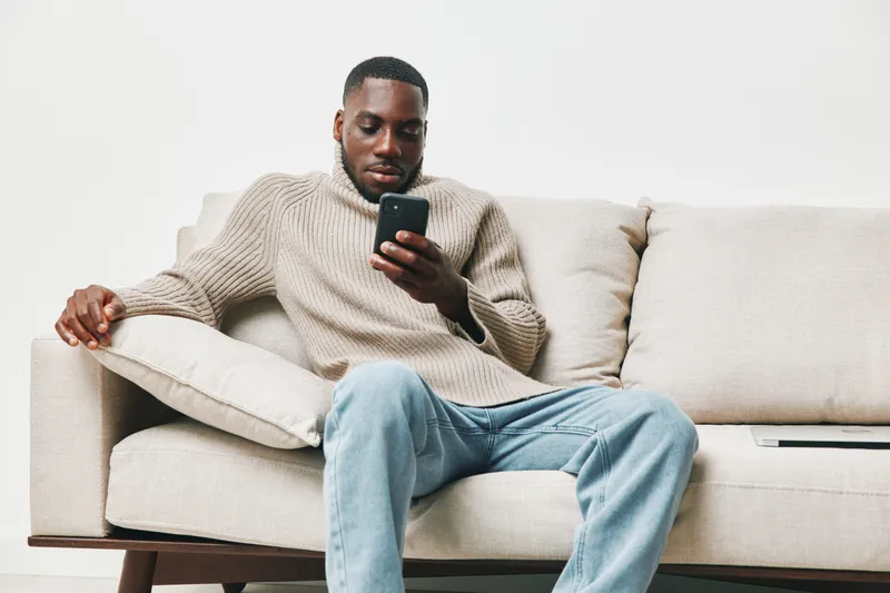 Young man casually dressed in a beige sweater and light jeans, sitting on a sofa while looking at his smartphone in a contemporary living room