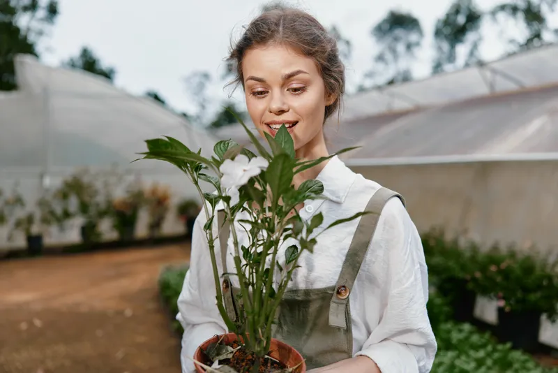 Young woman with light brown hair, wearing a casual outfit, joyfully holding a potted plant in a greenhouse, surrounded by greenery and natural light
