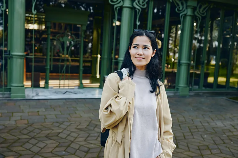 Woman in a beige coat and soft sweater stands outdoors by a green building, casual fashion vibe and backpack ready for urban exploration and daily errands