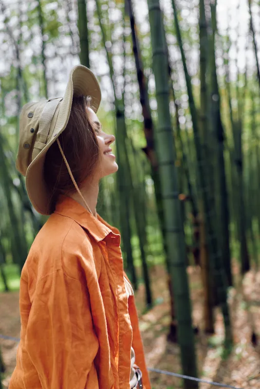 Woman profile with hat smiling in bamboo forest, orange jacket and relaxed pose, enjoying nature and peaceful outdoor walk among tall green stalks and filtered sunlight.