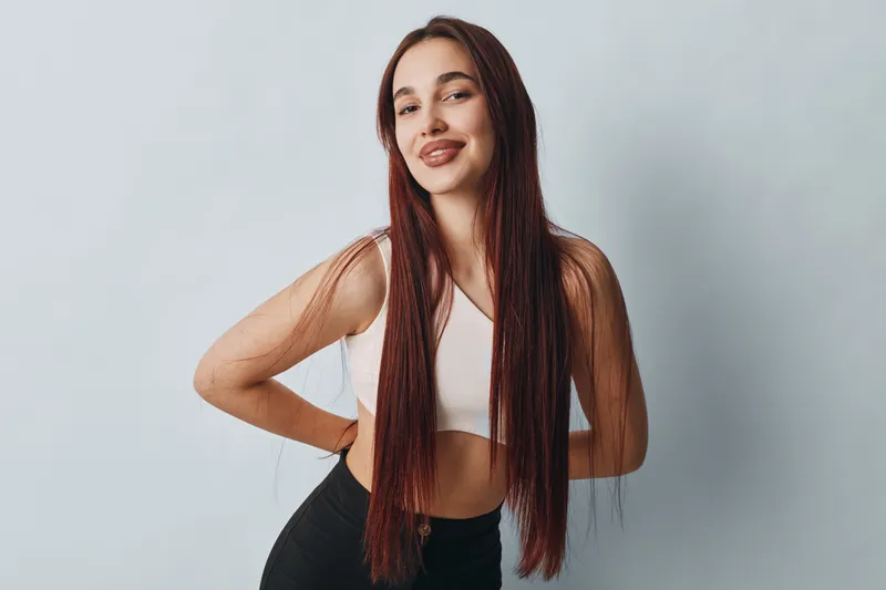 Young woman with long straight hair posing with hands on hips and smiling in white top and black pants against plain light background in studio portrait.