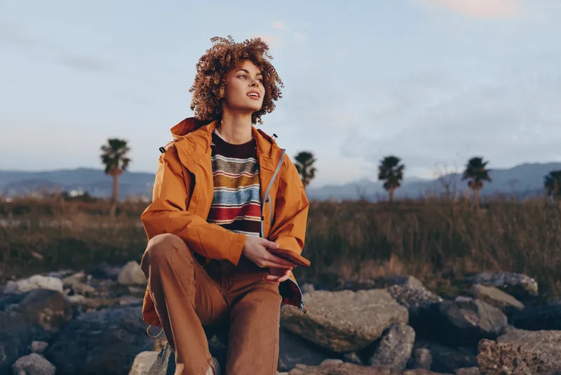 Woman lifestyle outdoor portrait, curly haired woman sitting on rocks in desert landscape, wearing orange jacket and striped shirt, relaxed calm mood at sunset, soft natural light